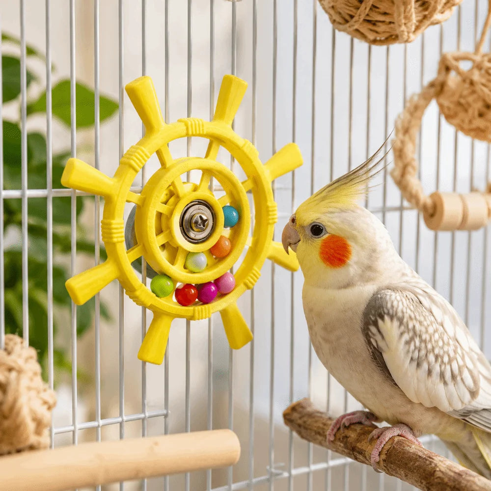 A cockatiel perched on a wooden branch inside a bird cage, interacting with a yellow ship's wheel bird toy that contains small colorful beads.