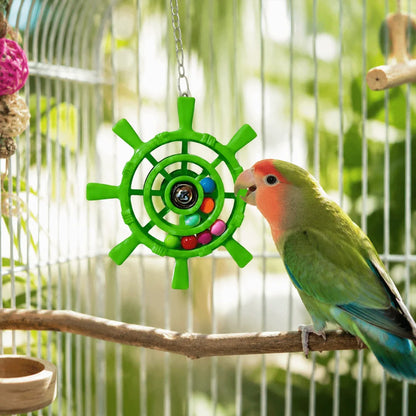 A green parrot with a peach face perched on a wooden branch inside a bird cage, interacting with a bright green ship wheel hanging toy that contains small colorful beads.