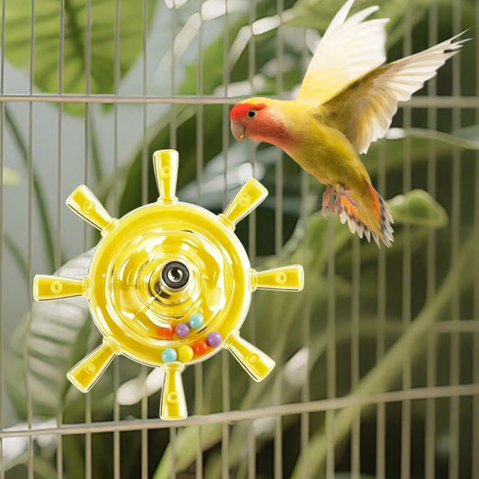 A small yellow lovebird with a red face and white wings flies in front of a birdcage while interacting with a bright yellow ship-wheel shaped toy that has colorful beads inside.