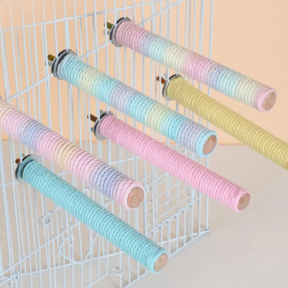 Six pastel rope bird perches wrapped in pastel rainbow-colored rope, mounted on a white wire cage against a light peach background.