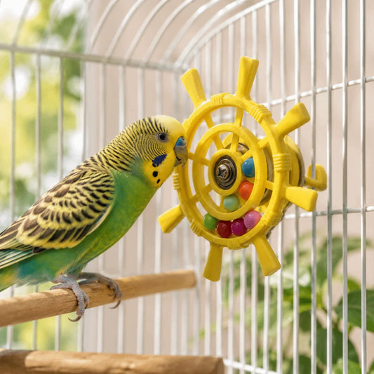 A green and yellow budgerigar perched on a wooden bar inside a white wire bird cage, interacting with a yellow ship's wheel toy filled with colorful beads.