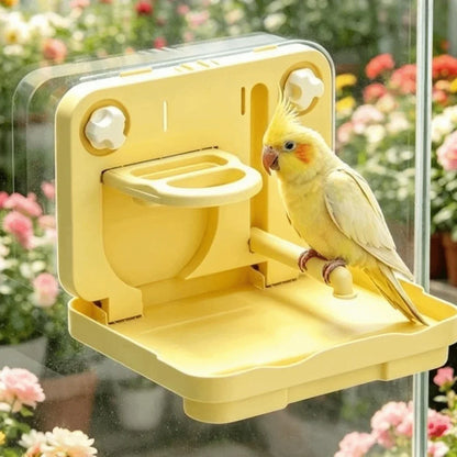 A yellow bird feeder mounted on a clear window, with a small yellow cockatiel perched on the feeder. The feeder has a hinged tray, a small bowl, and two white adjustment knobs on the back, set against a blurred background of pink and green flowers.