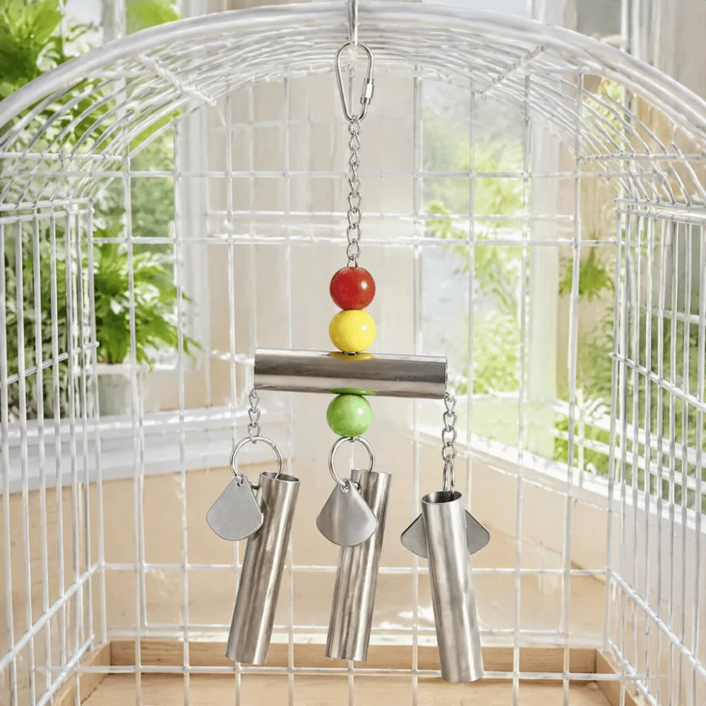A white wire bird cage with a hanging metal toy inside, featuring a horizontal bar, colorful beads in red, yellow, and green, and three hanging silver metal tubes.