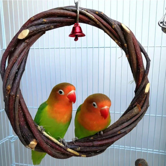 A bird toy hanging wreath made of dark brown vine branches, with a small red bell at the top and two lovebirds perched inside the circular wreath, set against a white wire bird cage.