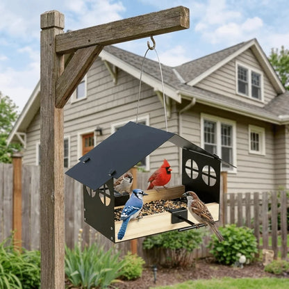 A house-mounted bird feeder hanging from a wooden post in a backyard, with several birds perched on the feeder. The feeder has a black metal frame with a slanted roof and a wooden tray below, set against a house and fence background.